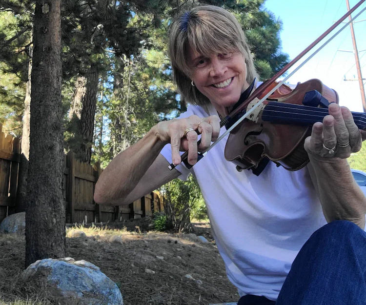 A smiling woman outdoors in a wooded setting, holding and playing a brown violin with a bow. She is wearing a white t-shirt and blue jeans, with tall pine trees and a blue sky in the background.